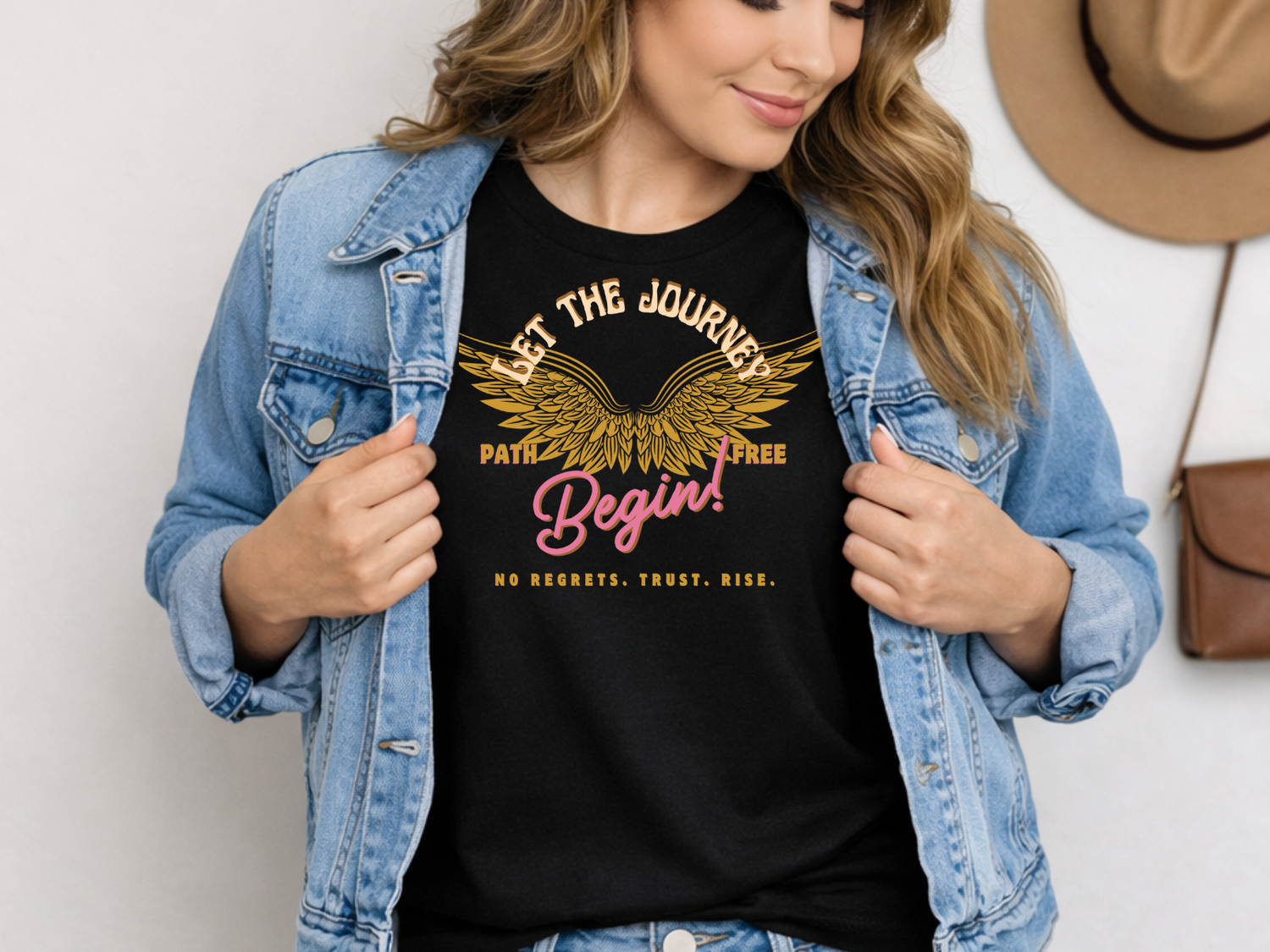 Woman wearing a black t-shirt with a motivational quote, standing against a light-colored wall with a hat and bag in the background.